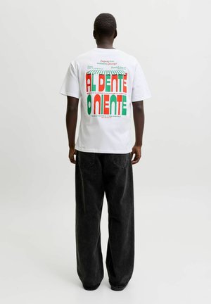 Personne portant un T-shirt blanc avec un grand texte rouge et vert « AL DENTE » et un pantalon noir, debout, dos à un fond uni.