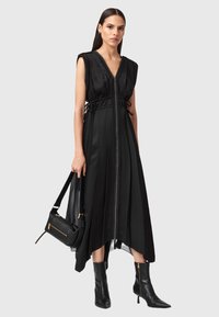Vestito nero senza maniche con scollatura a V e zip davanti, caratterizzato da una vita stretta e un orlo fluido. Indossato con stivali neri alla caviglia e una borsa a spalla.