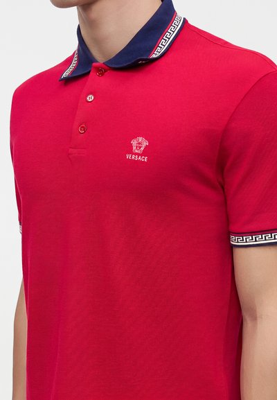 Άνδρας που φοράει κόκκινο polo Versace με ναυτικό γιακά και άκρες μανικιών με λευκό ελληνικό κλειδί σχέδιο περιθωρίου.