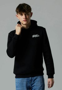 Giovane uomo che indossa una felpa nera con testo bianco e jeans blu, mentre tiene il colletto della felpa, su uno sfondo grigio neutro.