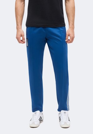 Mężczyzna w niebieskich spodniach dresowych Adidas z białymi paskami i białych butach sportowych Adidas, stojący na jednolitym tle.