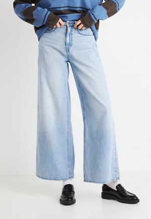 Vaqueros de pierna ancha en azul claro, con cintura alta, diseño de cinco bolsillos y dobladillo deshilachado. Combinados con mocasines negros y calcetines blancos.