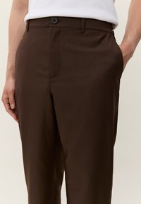 Oseba, oblečena v temno rjave hlače z gumbom in zadrgo ter stranskimi žepek, skupaj z belim brezrokavnikom.