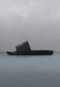 Sandale noire à enfiler avec une large sangle et un logo blanc discret sur une surface grise lisse et un fond gris clair.
