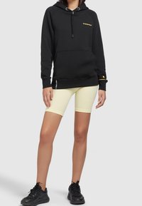 Czarna Bluza z kapturem z żółtym logo, kieszeń z przodu, kaptur ze sznurkiem; zestawiona z żółtymi szortami rowerowymi i czarnymi trampkami, stojąca na neutralnym tle.