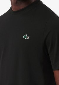 Svart crew-neck t-shirt med en liten grön och vit krokodil-logga på vänster bröst, bärs av en person med mörk hud.