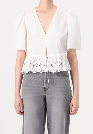 Femme portant un chemisier blanc à manches courtes avec une bordure en dentelle ajourée et des boutons, associé à un jean gris taille haute.