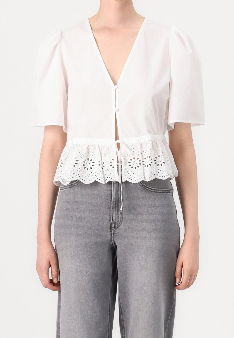 Femme portant un chemisier blanc à manches courtes avec une bordure en dentelle ajourée et des boutons, associé à un jean gris taille haute.