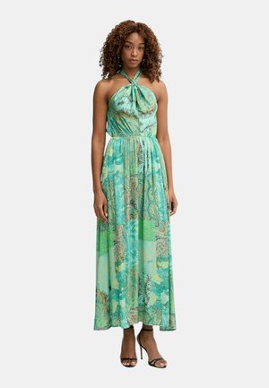 Donna con capelli ricci che indossa un abito lungo a collo halter verde fantasia e sandali neri con tacco alto, in piedi rivolta verso il davanti.