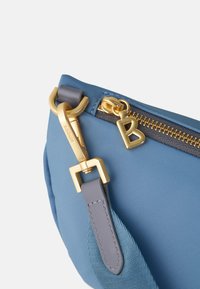 Borsa in nylon blu con hardware dorato, zip con charm e tracolla grigia testurizzata. Le forme includono un corpo arrotondato e dettagli piatti.