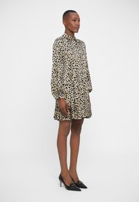 Vestido de leopardo en beige y negro, con mangas largas, un cuello abotonado y una falda acampanada. Combinado con tacones negros de puntera.