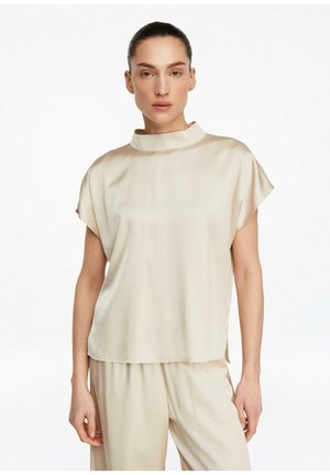 Frau mit dunklen Haaren in einem glatten Dutt, die eine cremefarbene Kurzarmbluse und passende weite Hosen vor weißem Hintergrund trägt.