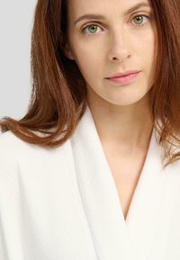 Ropa blanca texturizada con cuello de chal y un brillo sutil, que presenta un lazo gris claro en la cintura. Tejido suave y liso.