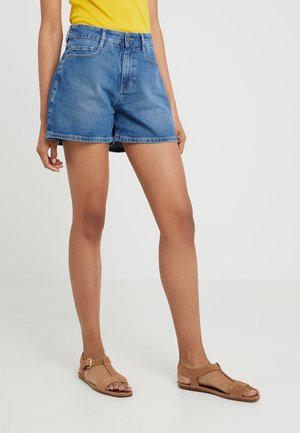 Person iført blå denimshorts, en gul top og lysebrune flade sandaler, stående mod en ensfarvet lys grå baggrund.