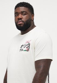 Beige t-shirt met korte mouwen featuring een bloemen- en tekstgrafiek op de linkerborst, gemaakt van zachte katoenstof. Standaard ontwerp met ronde hals.