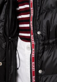 Chaqueta acolchada negra con textura brillante, cinta con el logo rojo de Levi's en la cremallera, y una camiseta de rayas color burdeos y blanca por debajo.