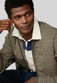 Brown checkered bomber jacket with a white collar over a blue and white striped polo shirt, featuring a textured fabric and zipper closure.