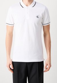 Vit polo t-shirt i bomull, med svart kant på kragen och ärmarna, samt en liten broderad logotyp på bröstet.