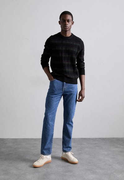 Pull noir avec un motif à carreaux et des accents verts et violets, associé à un jean bleu clair et des baskets beiges. Coupe simple et moderne.