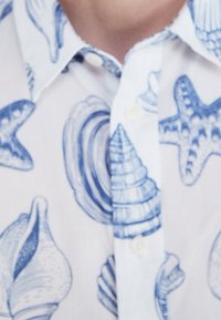 Košile s krátkým rukávem s bílým pozadím a ilustracemi modrých mušlí a hvězdic, s knoflíkovým límcem z jemně texturované látky.
