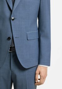 Veste de costume bleue avec boutons noirs, rabat de poche, pantalon assorti, manchette blanche de chemise, cravate sombre à motif et une main détendue le long du corps.