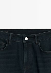 Gros plan sur la ceinture d'un jean denim bleu foncé avec bouton, passants de ceinture, poches avant et coutures visibles sur un fond blanc uni.