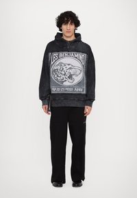 Haut de survêtement noir surdimensionné avec un imprimé graphique de tigre et le texte "LES BENJAMINS". Porté avec un pantalon cargo noir, présentant une texture lisse et une coupe décontractée.
