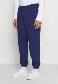 Mörkblå mjukisbyxor med elastisk midja och mudd vid bensluten, med en liten logotypdetalj på låret. Kombinerade med vita sneakers.