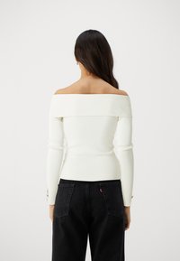 Schulterfreies Langarmoberteil aus weißem Rippenstoff, figurbetontes Design mit strukturierter Oberfläche, kombiniert mit dunklen hoch taillierten Jeans.