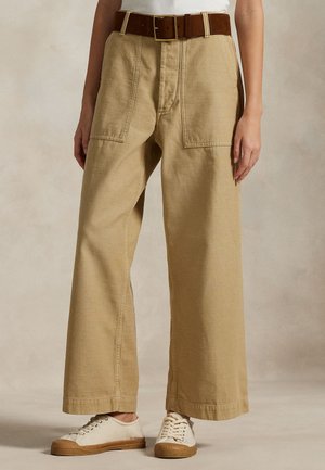 Γυναίκα που φοράει μπεζ παντελόνες φαρδιές με μεγάλες τσέπες μπροστά, καφέ ζώνη, λευκά αθλητικά παπούτσια και λευκή μπλούζα που είναι μέσα στην μέση.