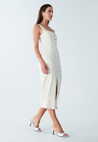 Robe midi blanche avec encolure carrée, boutons dorés sur le devant, fente latérale et bretelles fines. Associée à des escarpins blancs pointus sans lacets.