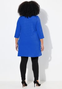Blå tunika med trekvartslånga ärmar, parat med svarta leggings och högklackade skor, sedd bakifrån.