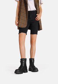 Shorts in denim neri con bordi sfilacciati, un gilet scozzese marrone e stivali neri in pelle con lacci spessi, dotati di lati testurizzati e dettagli in metallo.