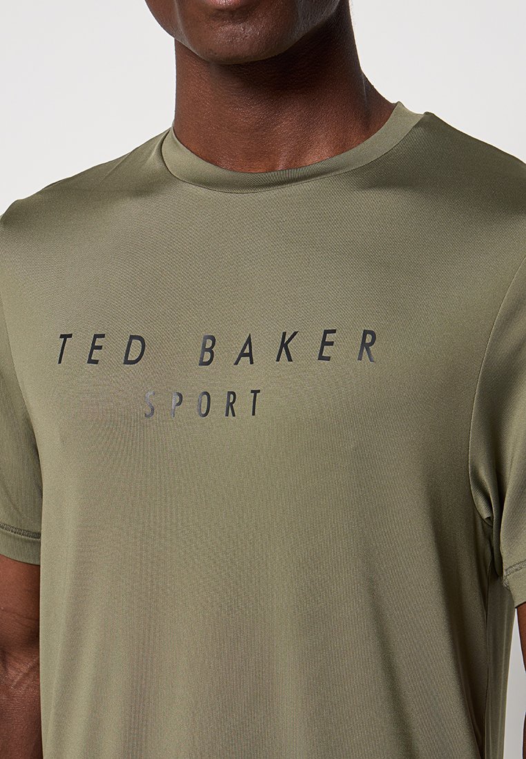 Persona de piel oscura vistiendo una camiseta deportiva Ted Baker de color verde oliva, cortada en el cuello y a la mitad del brazo, fondo liso.
