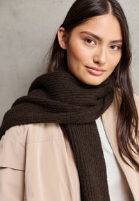 Écharpe tricotée marron foncé avec une texture côtelée, enroulée autour du cou, associée à une veste zippée beige clair. Fini en tissu lisse.
