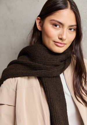 Dunkelbrauner gestrickter Schal mit geripptem Muster, um den Hals geschlungen, kombiniert mit einer hellbeige Jacke mit Reißverschluss. Sanfter Stoffabschluss.