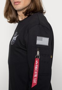 Sudadera negra con gráfico del logo de la NASA, parche de la bandera estadounidense, bolsillo con cremallera y etiqueta roja "REMOVE BEFORE FLIGHT". Textura suave.