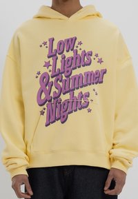 Gelbe Kapuzenjacke mit weiter Passform, die mit auffälligem lila Text "Low Lights & Summer Nights" und Sternen-Grafiken versehen ist. Material aus Baumwollmischung.