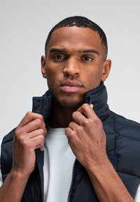 Schwarze Steppweste mit hohem Kragen, ausgestattet mit einem Reißverschluss und horizontalen Nähten, getragen über einem hellgrauen Hemd. Hände greifen den Kragen.