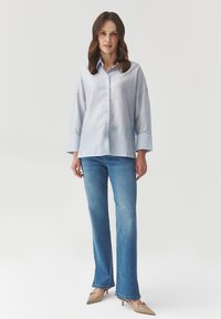 Camisa de botones a rayas azul claro con cuello, combinada con jeans azules de pierna acampanada y tacones beige. Tejido suave con un ajuste relajado.