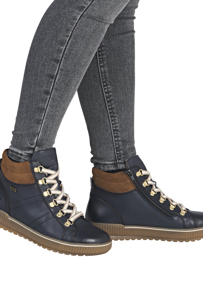 Baskets montantes en cuir bleu marine avec des accents marron, des garnitures dorées, des lacets blancs et une semelle en caoutchouc texturée. Fermeture éclair sur le côté.