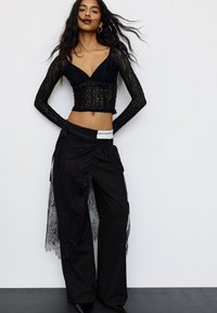 Femeie cu părul lung și închis la culoare, îmbrăcată cu un top scurt cu mâneci lungi din dantelă neagră, pantaloni largi negri și o fustă din dantelă neagră peste aceștia.