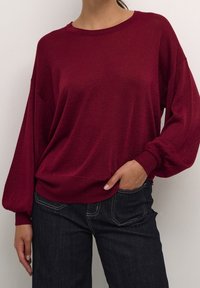Pull-over bordeaux à coupe décontractée, poignets côtelés et texture en maille légère. Porté avec un jean en denim foncé.