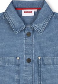 Chemise en denim bleu clair, avec un col évasé, deux poches sur le devant, et des boutons noirs avec logo. Tissu texturé avec des rayures verticales.