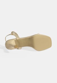Goldene, metallisch glänzende Sandale mit hohen Absätzen, quadratischem Absatz und verstellbarem Knöchelriemen. Glattes Material und strukturierte Sohle für besseren Halt.