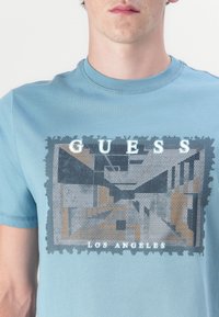 Šviesiai mėlynas medvilnės marškinėliai su grafiniu spaudiniu, vaizduojančiu pastato sceną, su žodžiu "GUESS" viršuje ir "LOS ANGELES" apačioje.