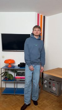 Jaunas vyras mėlynoje hoody ir džinsuose stovi kambaryje su prie sienos pritvirtintu televizoriumi, oranžine lempa, kamera, knygomis ir metaliniu dėklu su lipdukais.