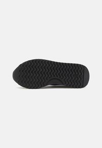 Semelle de chaussure en caoutchouc noir avec un motif de marche texturé et ondulé. Présente une empreinte de logo circulaire près du talon.
