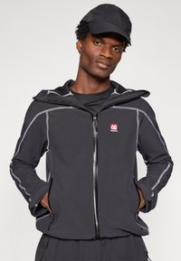 Jaqueta preta com fecho zip e costura cinza, com capuz. Logotipo destacado no lado esquerdo do peito. Tecido suave com bolsos laterais, combinada com um boné preto.