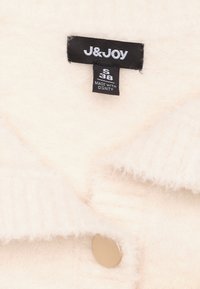 Crème-kleurige trui met een zachte, fluweelachtige textuur, geribbelde kraag en een ronde gouden knop. Etiket zichtbaar met merknaam "J&JOY" en maat "S 38."